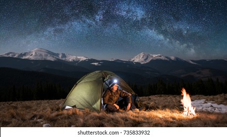 Male Tourist Have A Rest In His Camping In The Mountains At Night. Man With A Headlamp Sitting Near Campfire And Tent Under Beautiful Night Sky Full Of Stars And Milky Way.