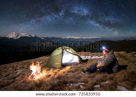 Male tourist have a rest in his camp at night. Guy with a headlamp sitting near campfire and tent under beautiful sky full of stars and milky way. On the background snow-covered mountains