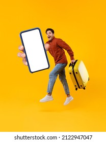 Male Tourist Jumping Holding Huge Smartphone And Suitcase Showing Blank Cellphone Screen To Camera Advertising Great Travel Application Posing In Mid Air On Yellow Background In Studio. Vertical