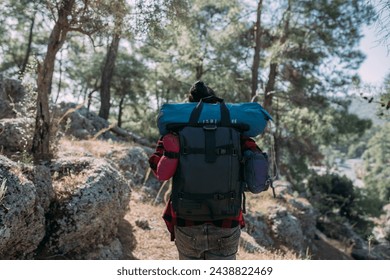 A male tourist with a large backpack and equipment on a mountain trail among the trees.  A young guy is walking alone along a hiking trail in the mountains with a backpack, a tent and a sleeping bag - Powered by Shutterstock