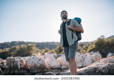 A male tourist with a large backpack and hiking gear on a rocky seashore.  A young guy walks along the coastal large stones on a hiking trip along the ocean shore - Powered by Shutterstock