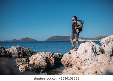 A male tourist with a large backpack and hiking gear on a rocky seashore.  A young guy walks along the coastal large stones on a hiking trip along the ocean shore - Powered by Shutterstock