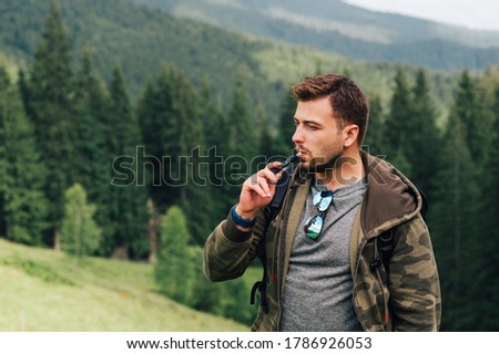 Male tourist uses an electronic cigarette with the evaporation of tobacco on a hike in the mountains, standing and resting. Bearded man smokes an electron cigarette in the mountains on a meadow.