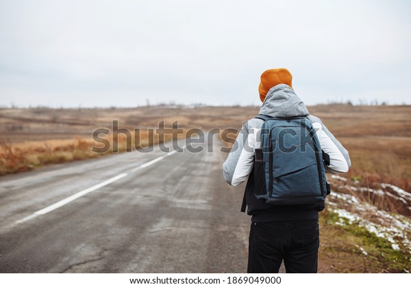 Male tourist walks on the side of an empty\
winter road in suburban area with a backpack. A man traveller\
wearing orange hat on a highway. All seasons tourism. Hitch-hiking,\
travelling concept.