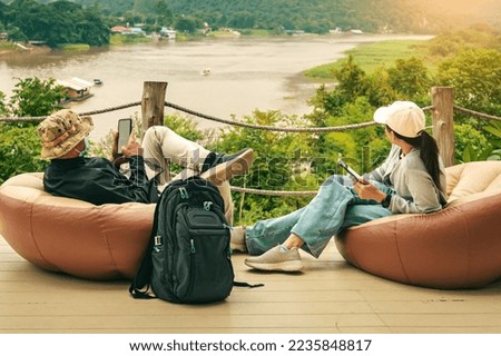 Male tourist wear hat with face mask and his wife use mobile phone and sit to relax on large cushions on balcony with mountains and river in background. Traveller in bag chair enjoy outdoor nature.