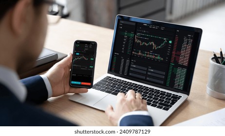 Male trader sits at desk hold smart phone with stock market data, using laptop display graph and chart, analyzing investment, check trading stocks information, view over shoulder. Growth, profit, apps - Powered by Shutterstock