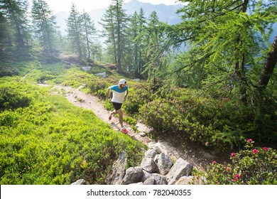 A Male Trail Runner Running Along A Trail In The Mountains, Through A Green Landscape With Trees And Plants In Spring. 