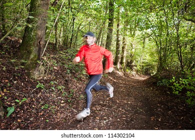 Male Trail Runner Running In The Forest On A Trail. Red Shirt And Black Pants. Summer Season. Slight Blur In Runner To Show Motion. Horizontal Composition.