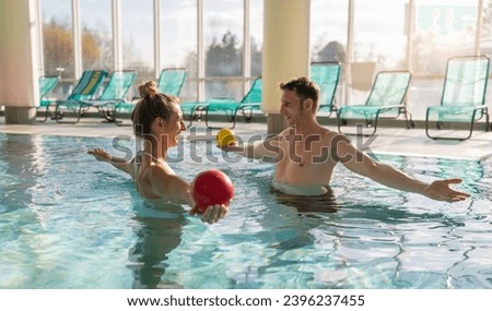 Male trainer conducting aquatic therapy with a female client using exercise balls in a sunny pool