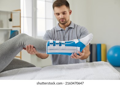 Male Traumatologist Provides Medical Care To Patient With Sprained Leg Ligament. Injured Leg Of Female Patient Wearing Blue And White Ankle Bandage With Adjustable Fracture Fixator Straps.