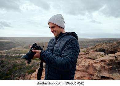 Male travel photographer checking photos on camera after shot standing on mountain wearing cap