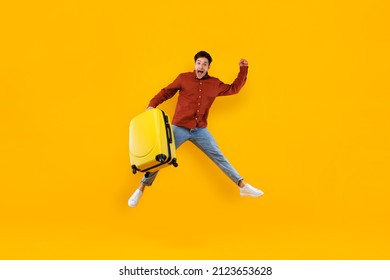 Male Traveler Jumping Holding Big Suitcase Looking At Camera Posing In Mid Air In Studio Over Yellow Background. Vacation, Finally. Man Celebrating New Journey Concept. Full Length