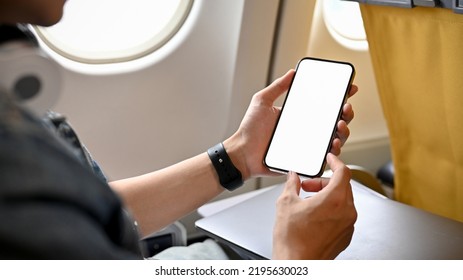 A male traveler passenger sits at the window seat in economy class, using his smartphone, holding a mobile phone white screen mockup. close-up image - Shutterstock ID 2195630023
