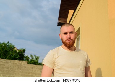 Male With Trendy Hairdo And Beard, Looks With Serious Expression, Has Thick Red Beard. Red-bearded Man On The Street In Summer. Male 28-35 Years Old. The Guy Against The Sky.