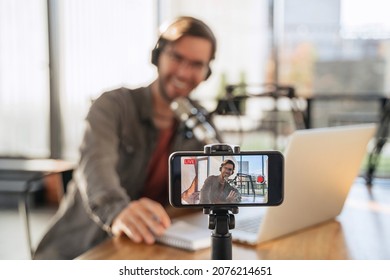 Male trendy podcast creator recording video podcast on smartphone. Selective focus on smartphone camera screen with man podcaster vlogger in headphones and glasses shooting live video for his channel
