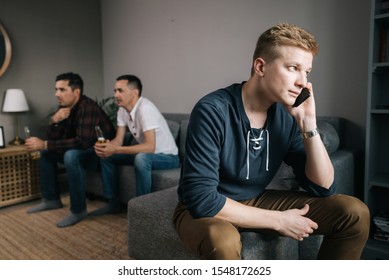 Male Trying To Talk On The Phone. Behind The Guy Back His Friends Are Watching TV. Group Of Friends Watching A Football Match On TV Against The Background Of Young Man Phone Conversation.