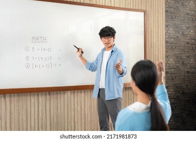 Male tutor standing in front of whiteboard is pointing to student for ask question and young students raising hands in the air to answer a question when learning in the classroom