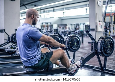 Male Using Rowing Machine At Fitness Club. Young Man Doing Exercises On Fitness Machine In Gym. Side View.dressed In Sportswear