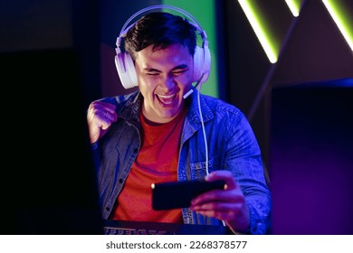 Male video game player cheering and celebrating as he streams a live broadcast of a game on his smartphone. Young man joining an online audience in watching a virtual gameplay on a mobile app.