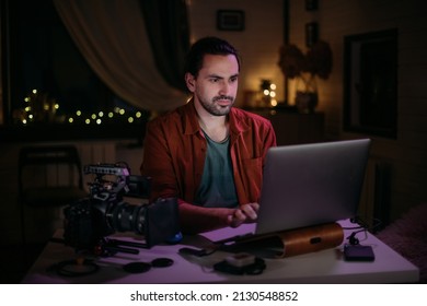 Male videographer at home workplace at table with laptop and camera. A young guy works, studies, makes content, mounts a video at home late at night, sits at a computer in a cozy room - Powered by Shutterstock