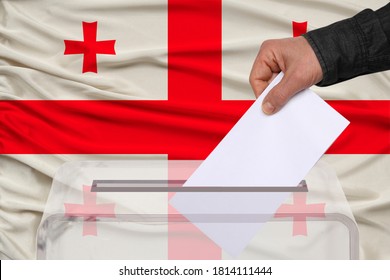 Male Voter Drops A Ballot In A Transparent Ballot Box Against The Background Of The National Flag Of Georgia, Concept Of State Elections, Referendum