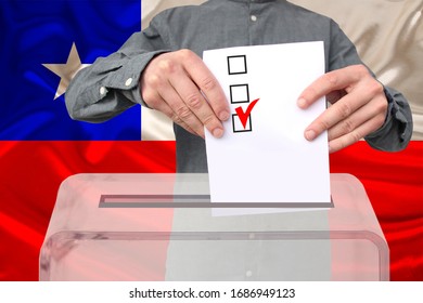 Male Voter Lowers The Ballot In A Transparent Ballot Box Against The Background Of The National Flag Of The Country Of Chile, Concept Of State Election, Referendum