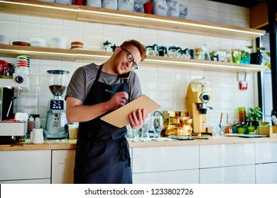 Male waiter in apron writing down order while talking on smartphone with client.Caucasian young entrepreneur calling on mobile phone to checking data standing near bar in own coffee shop - Powered by Shutterstock