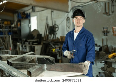 Male welder standing with a welding semi-automatic machine and a safety helmet in a metal machining workshop - Powered by Shutterstock