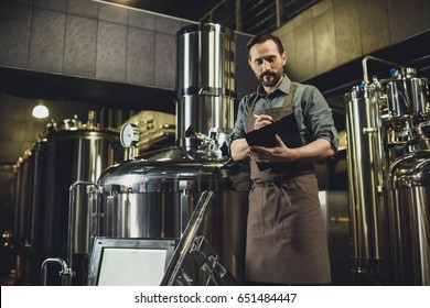 Male worker in apron inspecting industrial equipment and making notes at the brewery - Powered by Shutterstock