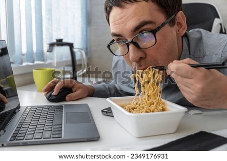 Male worker busy working with laptop, use chopsticks to hastily eat instant noodles during office lunch's break, because quick, tasty and cheap. fast food, unhealthy lifestyle.
