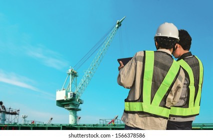 Male Worker At Construction Site With Hand Holding IPad Work Wearing Safety Helmet Hard Hat On Green Pipe Line And Crane In Project Site Background