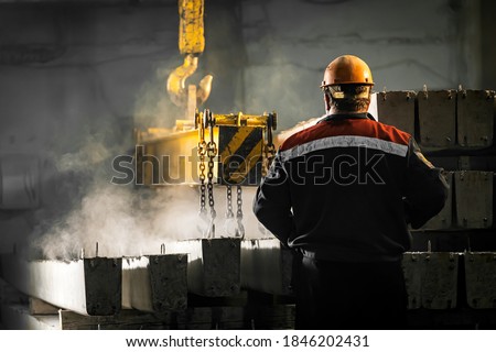 A male worker controls the production process in a factory as a crane moves a reinforced concrete product with holes. Reinforced concrete pillars secured with metal hooks and chains 