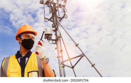 Male Worker Engineer Wearing A Mask, Helmet And Safety Goggles Works In The Field On A Telecommunication Tower. Supervise The Installation Of Radio Communication Electrical Equipment With Colleagues.