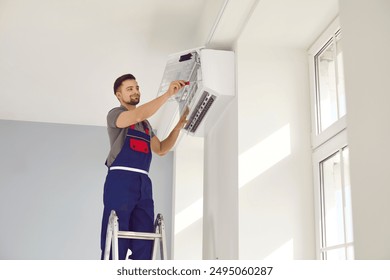 Male worker fixing air conditioning at home. Man in uniform standing on ladder stairs and installing new air conditioner on white wall in house. AC installation, maintenance, repair service concept