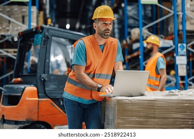 A male worker in a hard hat and safety vest is focusedly using a laptop atop stacked goods near a forklift inside a busy warehouse, conveying concentration and technological integration. - Powered by Shutterstock