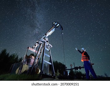 Male worker holding wrench and raising hand while standing near pump jack under beautiful night sky with stars. Petroleum engineer controlling work of oil pump rocker-machine at night in oil field.