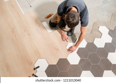 Male worker installing new wooden laminate flooring. The combination of wood panels of laminate and ceramic tiles in the form of honeycomb. Kitchen renovation. - Shutterstock ID 1211013040