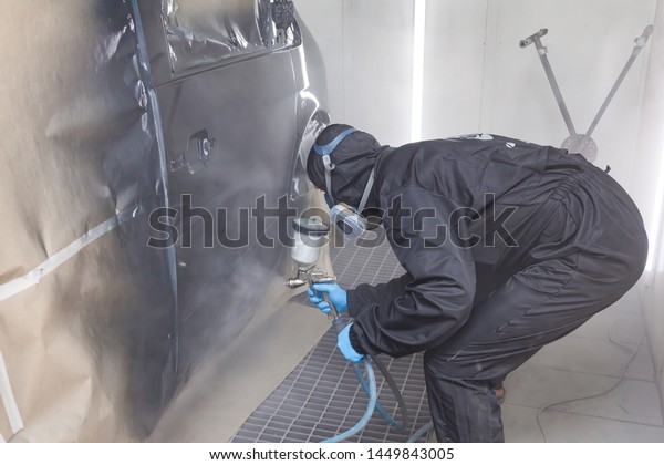 A male worker\
in jumpsuit and blue gloves paints with a spray gun a side part of\
the car body in black after being damaged at an accident. Auto\
service industry professions