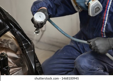 A Male Worker In Jumpsuit And Gloves Paints With A Spray Gun A Front Frame Part Of The Car Body In Black After Being Damaged At An Accident. Auto Service Industry Professions