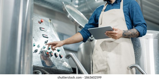 Male worker on beer factory, management and mobile app. Busy male employee or owner in apron with digital tablet controls equipment at brewery and push button, cropped, free space, panorama