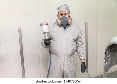 A Male Worker Painting A Car Is Standing In A Spray Booth In Protective Clothing With A Spray Gun In His Hands Pointing Forward And Looking Into The Camera