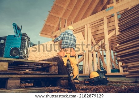 Male Worker Preparing to Start Work on the Wooden House Frame Building by Moving in the Plywood Plate, His Toolbox and Necessary Equipment. Construction Site in the Background. Industrial Theme.