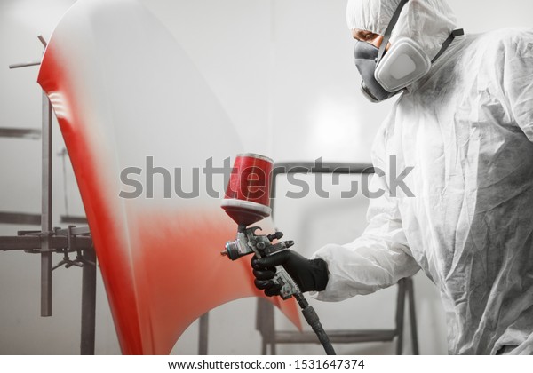 Male worker in protective clothes and\
mask painting hood of car using red spray\
paint.