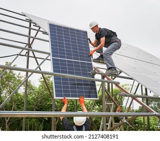 Male Worker In Safety Construction Helmet Giving Solar Module To Colleague. Two Men In Workwear Building Photovoltaic Solar Panel System. Concept Of Sustainable Energy And Solar Panel Installation.