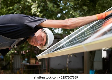 Male Worker In Safety Helmet Looking At Camera And Winking While Mounting Photovoltaic Solar Panel. Man Assembling Solar Modules For Generating Electricity Through Photovoltaic Effect.