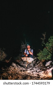 A Male Worker Is Sitting In A Wheelbarrow. Night View. Headlamp Illuminates The Trail. The Concept Of Heavy Utility And Construction Work In The Mountains On A Night Shift. Vertical Photo. Fun