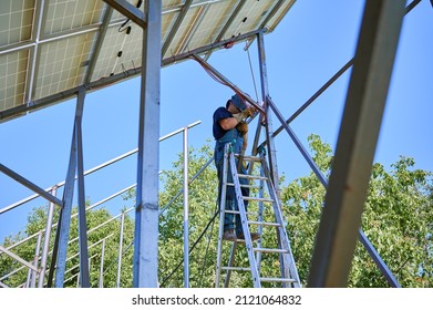Male Worker Standing On Ladder And Welding Metal Poles Of Photovoltaic Solar System. Man Wearing Safety Welding Mask While Mounting Support Structures For Solar Panels.