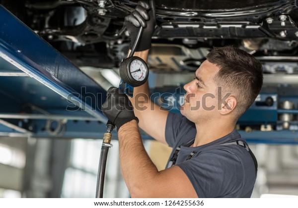 Male\
worker standing under car and checking pressure with meter in auto\
service. Serious mechanic in uniform and protective gloves in\
process of fixing vehicle. Concept of car\
repair.