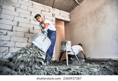 Male worker using shovel while shoveling sand-cement mix in building under construction. Man preparing floor screed material while standing near concrete screed mixer machine. - Powered by Shutterstock