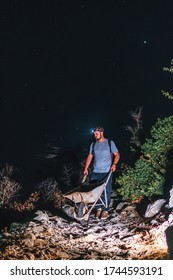 A Male Worker Is Walking With A Wheelbarrow. Night View. Headlamp Illuminates The Trail. The Concept Of Heavy Utility And Construction Work In The Mountains On A Night Shift. Vertical Photo